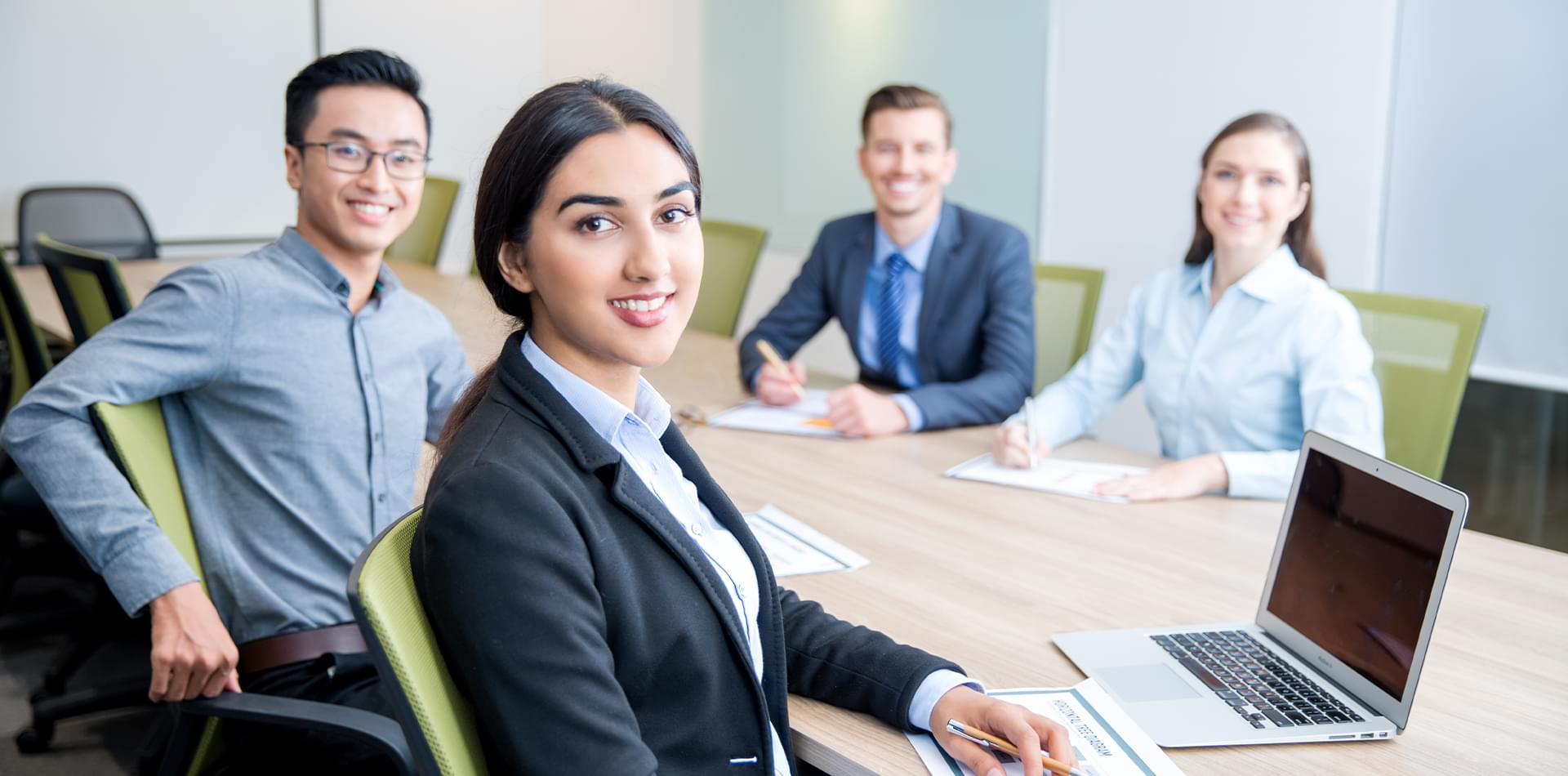A diverse group of professionals sit around a conference table in a modern meeting room. A woman in the foreground turns toward the camera with a confident smile while colleagues in business attire sit behind her with notebooks and a laptop, suggesting a collaborative team meeting or leadership discussion.